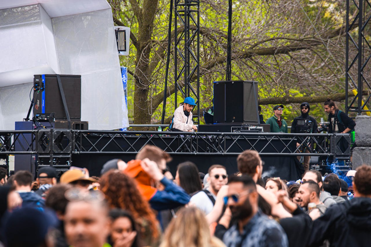 A DJ performs live on stage surrounded by a vibrant outdoor crowd, creating an energetic music festival atmosphere.